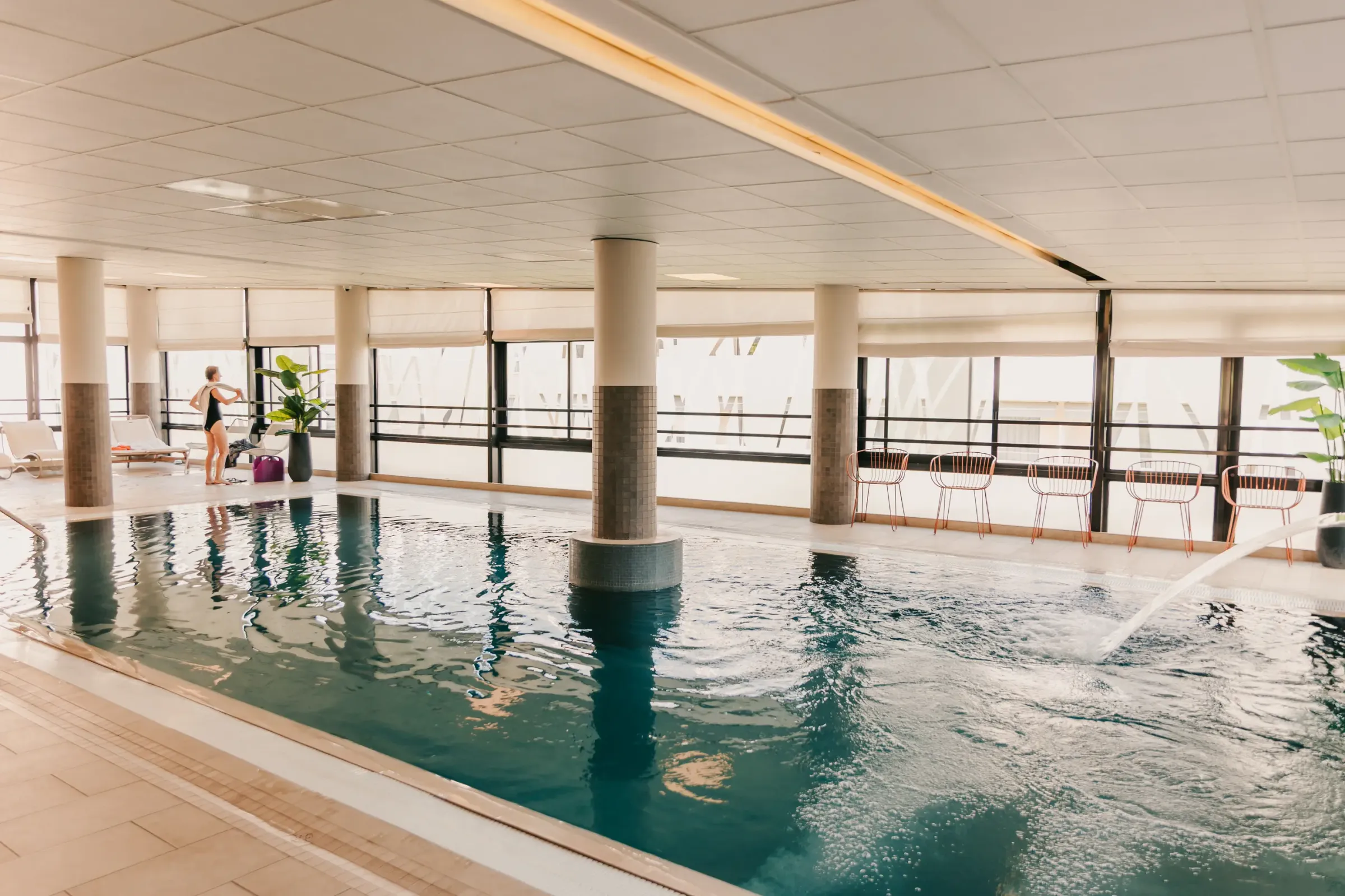Innenpool mit einer Person in Badebekleidung in der Nähe von Liegestühlen und Tischen, umgeben von großen Fenstern und Säulen