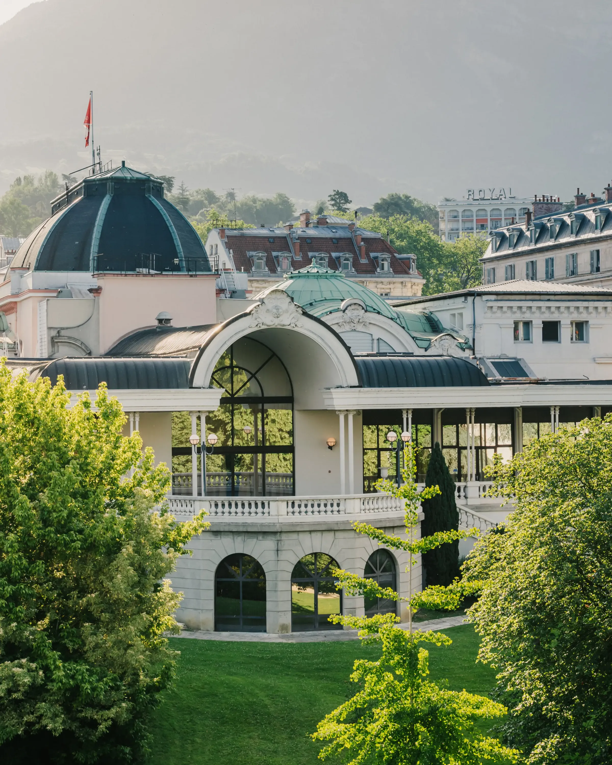 Un bâtiment avec une architecture classique et une grande verrière, entouré d'arbres et de pelouses, avec d'autres structures et un drapeau en arrière-plan
