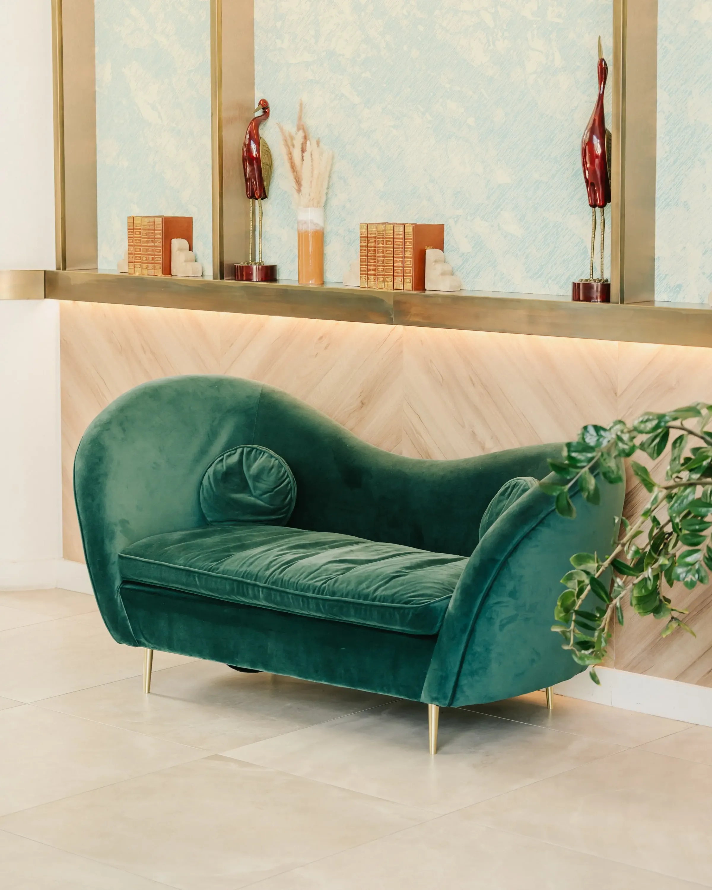 Un canapé vert en velours avec des pieds dorés est placé contre un mur en bois clair, sous une étagère décorée d'objets et de livres