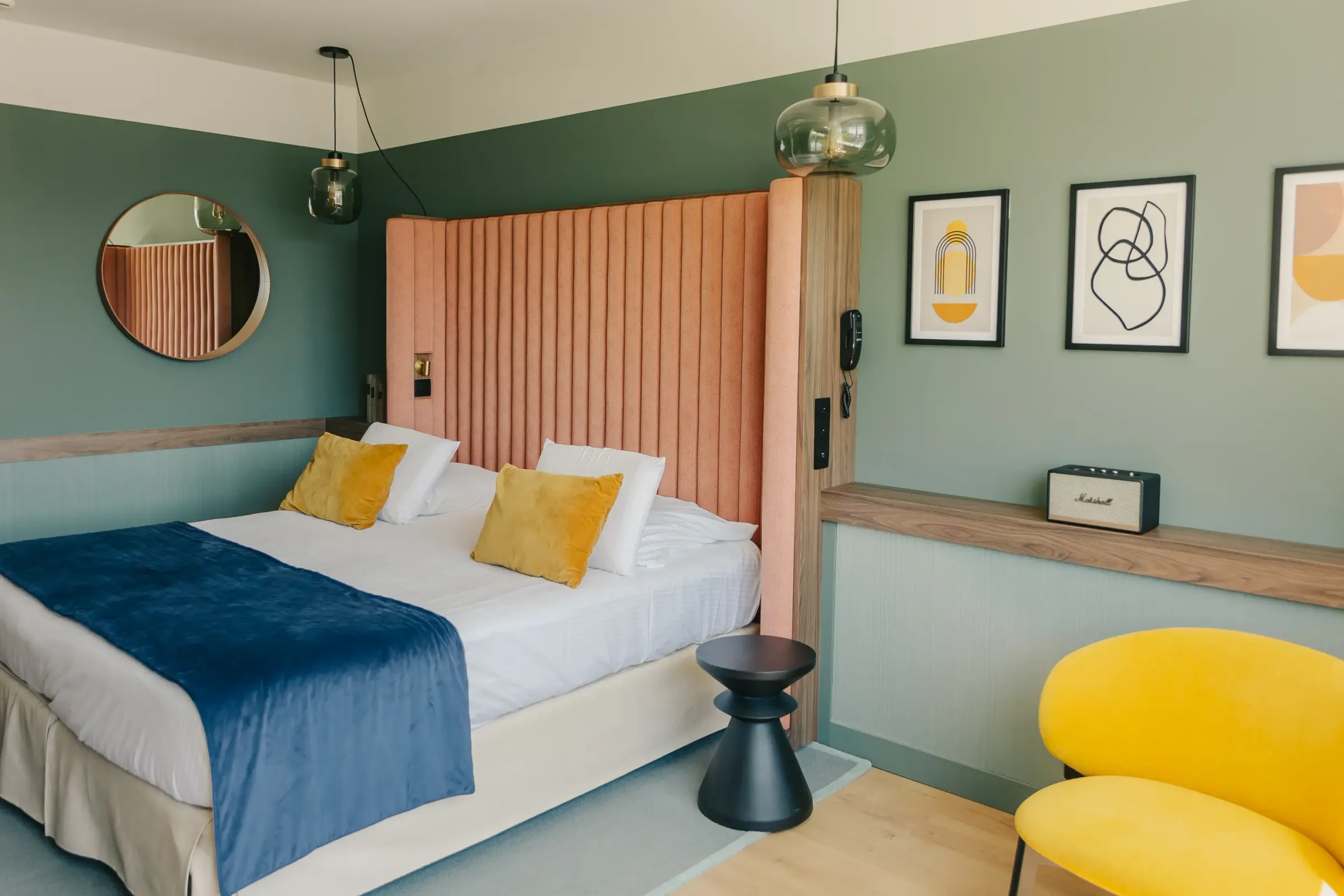 Chambre avec un lit blanc et bleu, des coussins jaunes, une tête de lit orange, une lampe murale, des tableaux aux murs et un fauteuil jaune