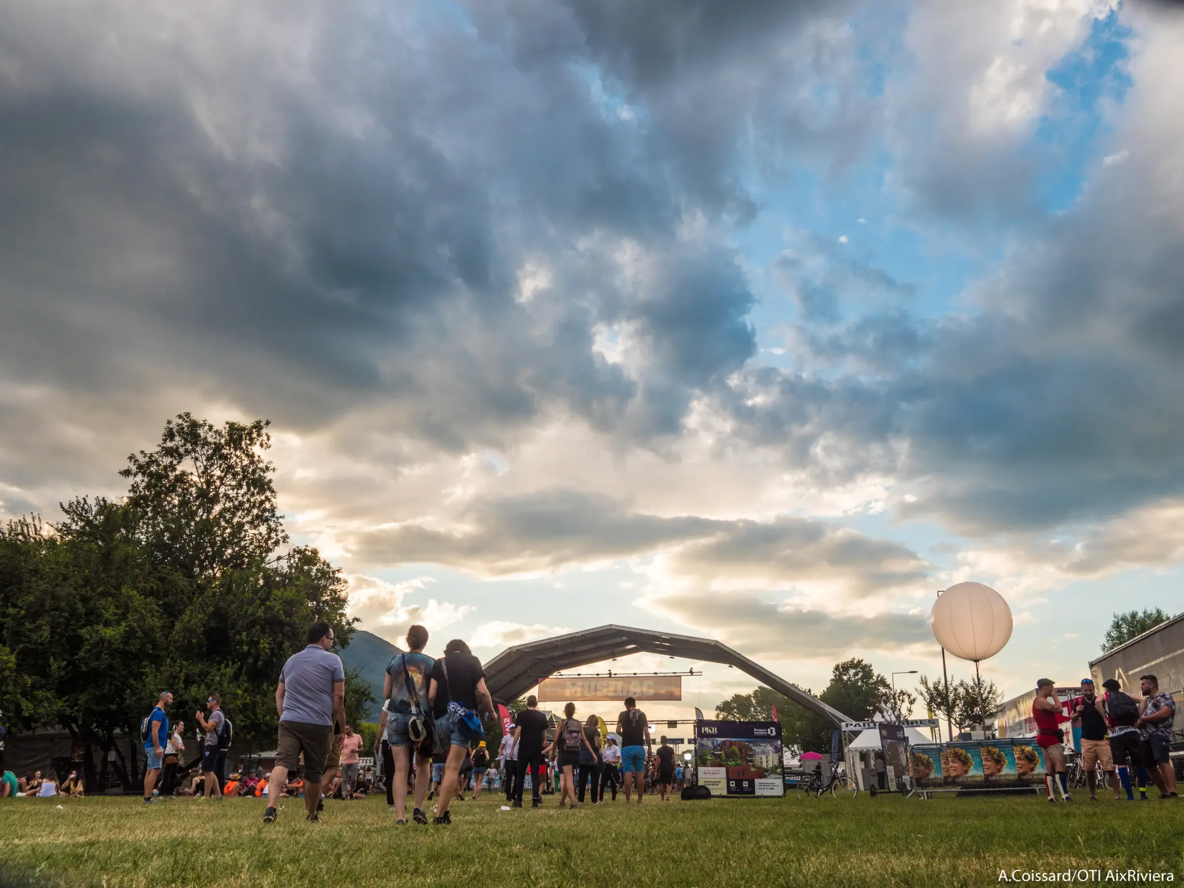 Un groupe de personnes se tient sur une pelouse devant une scène de concert sous un ciel nuageux avec un ballon blanc en forme de poire à proximité