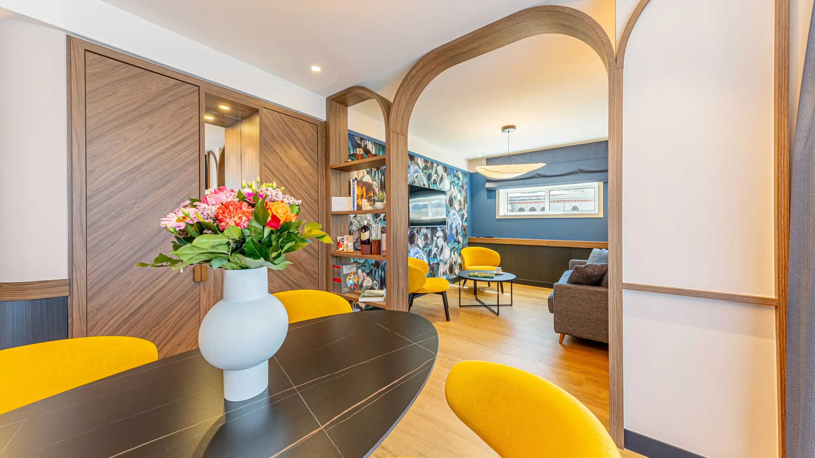 Moderner Salon mit schwarzem Tisch, gelben Stühlen, Blumenvasen und Bibliothek in einem Nebenraum