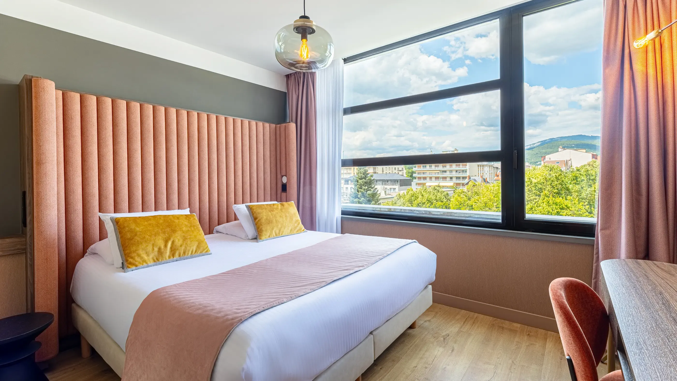 Chambre avec un lit double aux draps blancs et une couverture rose, deux coussins jaunes, une tête de lit orange et une vue sur des bâtiments et des arbres par une grande fenêtre