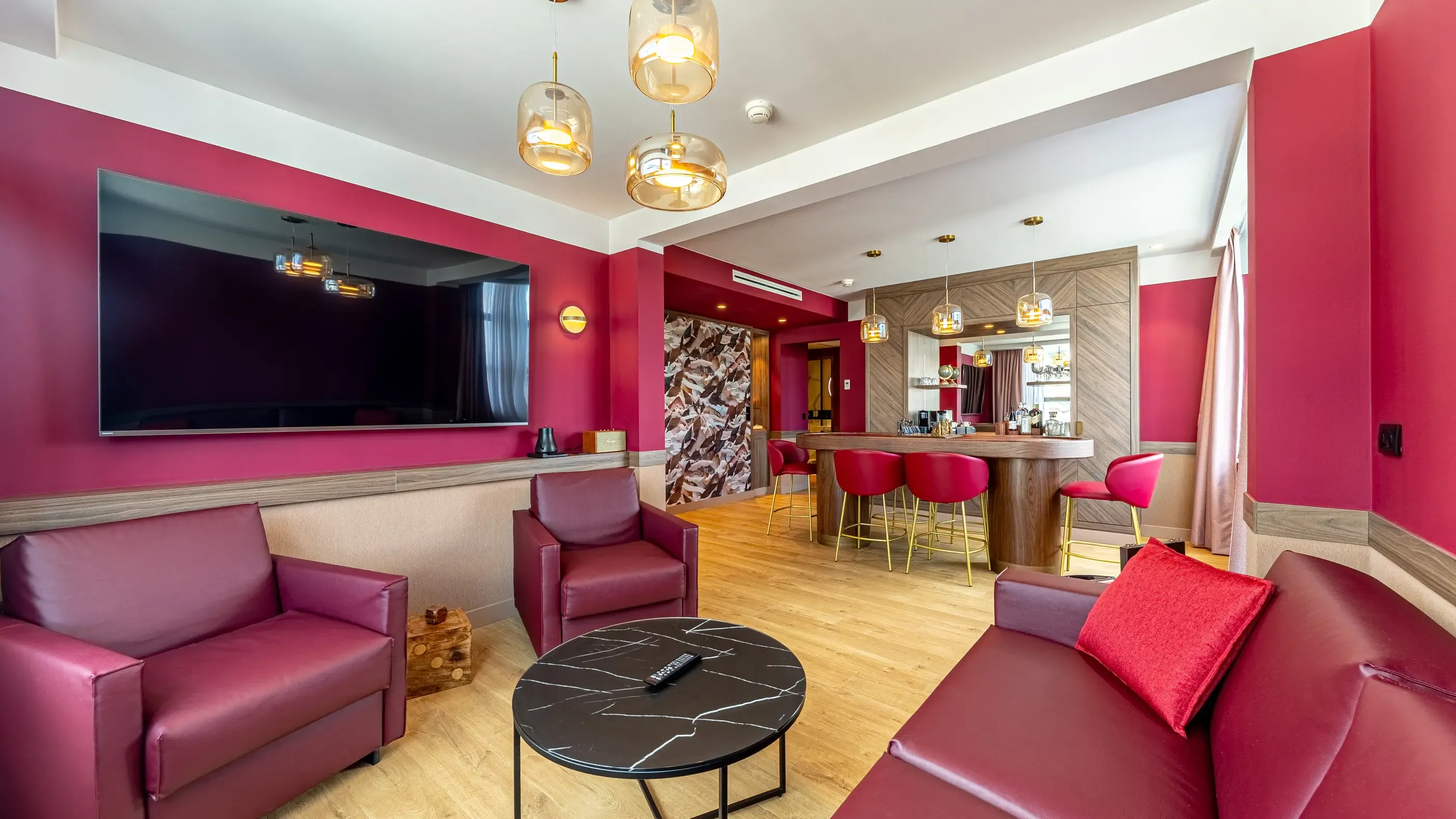 Salon avec canapés et fauteuils rouges, table basse en marbre noir, bar avec tabourets et écran mural