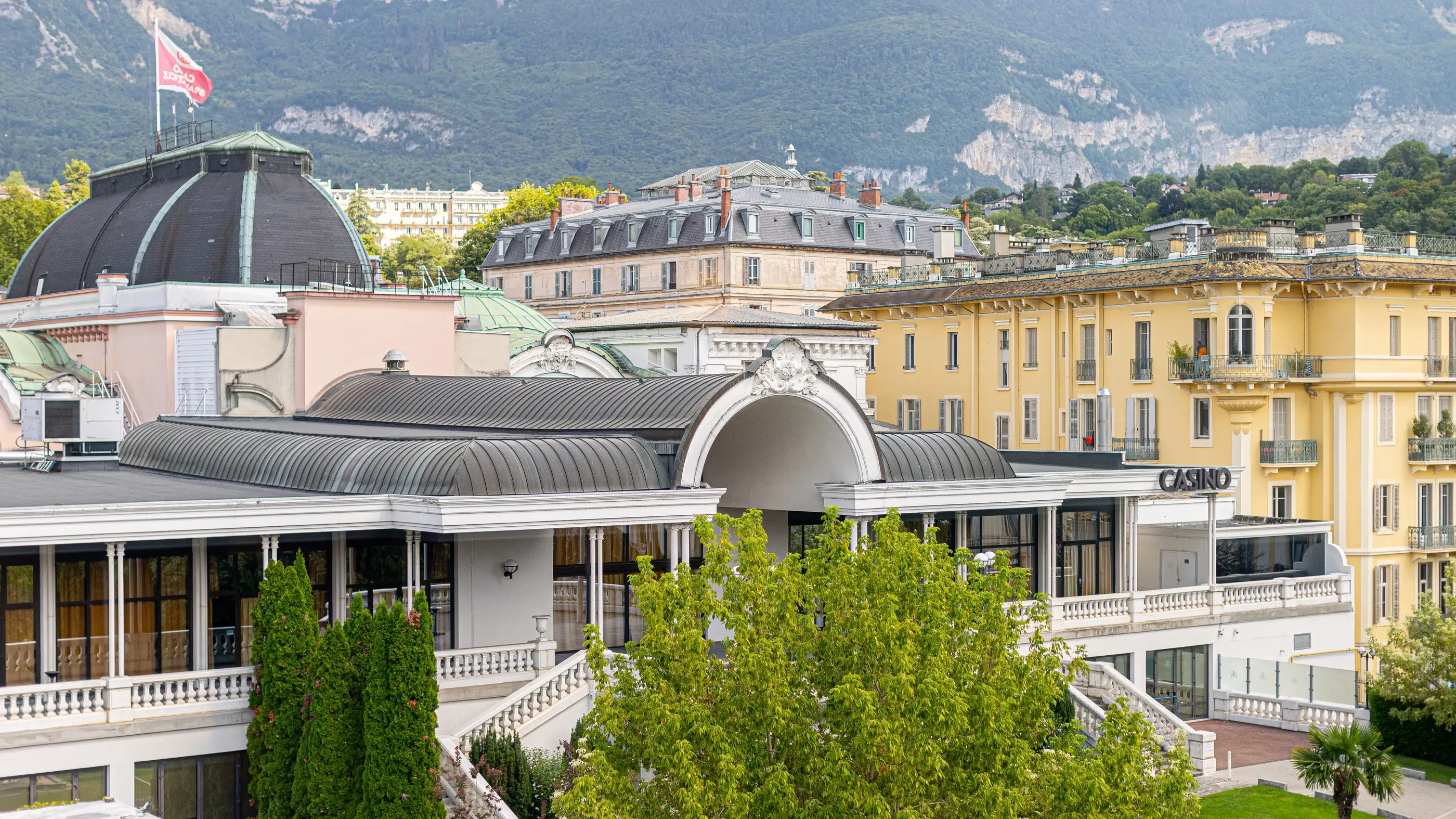 Un casino avec une architecture classique et des toits métalliques, entouré de bâtiments historiques et de montagnes en arrière-plan