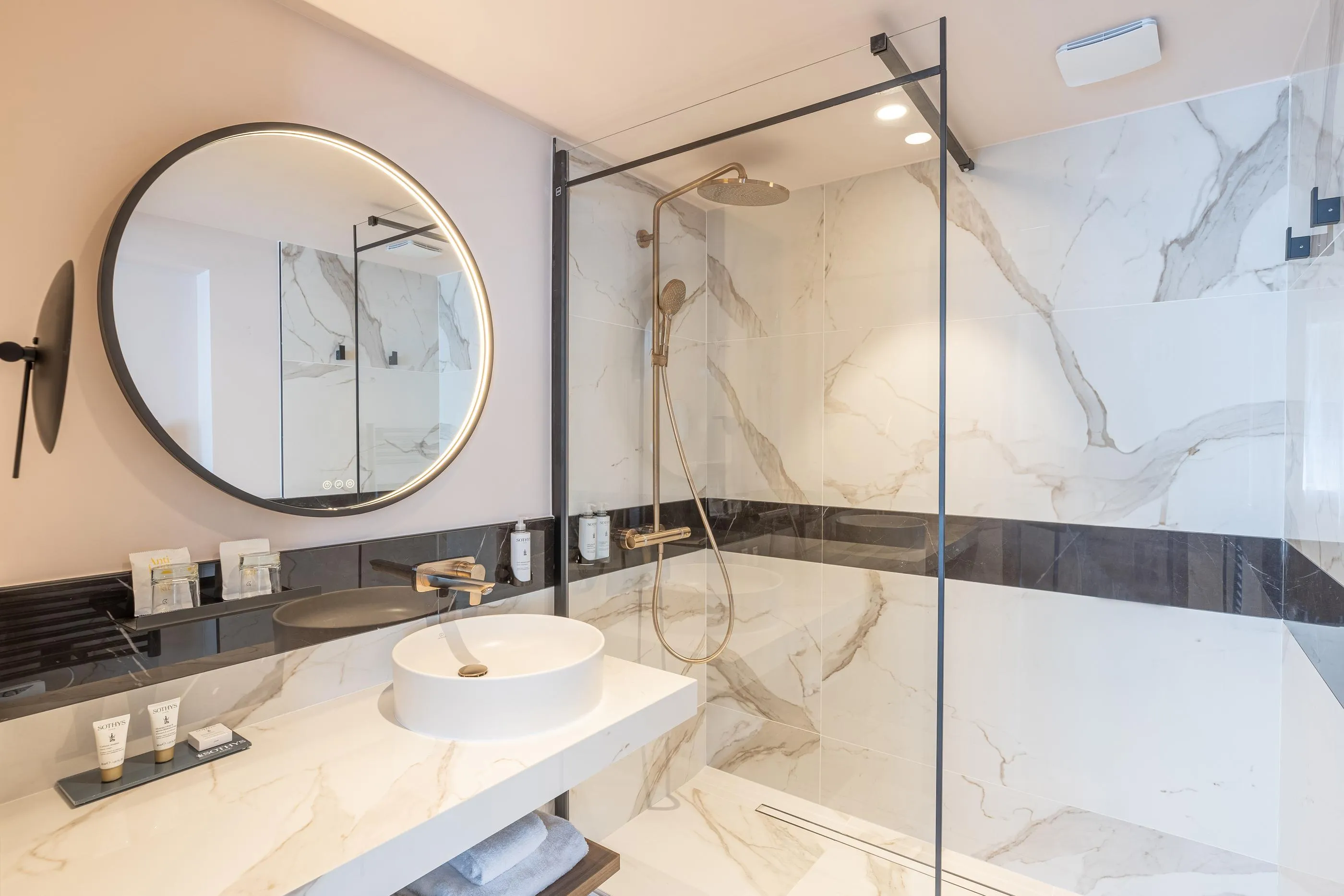 Salle de bain moderne avec lavabo, miroir rond et douche à effet pluie derrière une paroi en verre