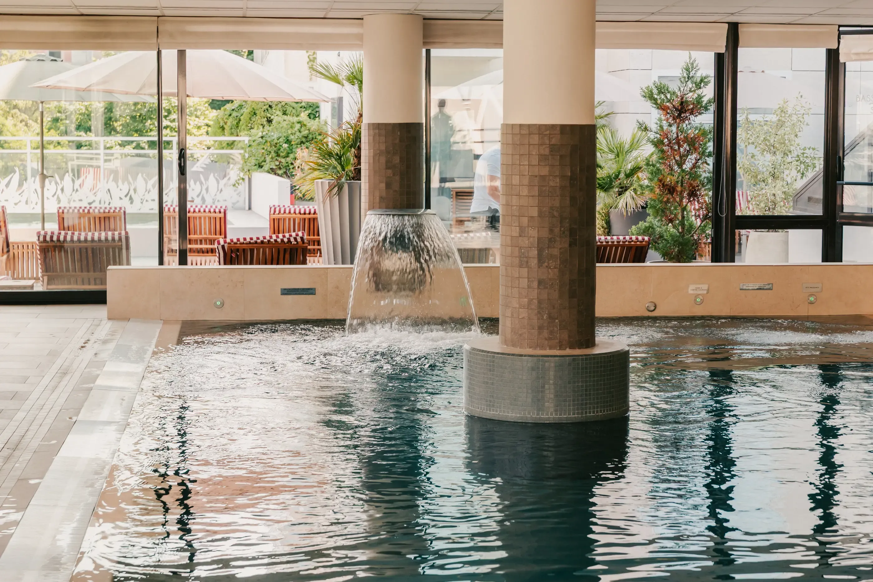 Piscine intérieure avec une fontaine centrale et des tables et chaises en terrasse visibles à travers les grandes baies vitrées