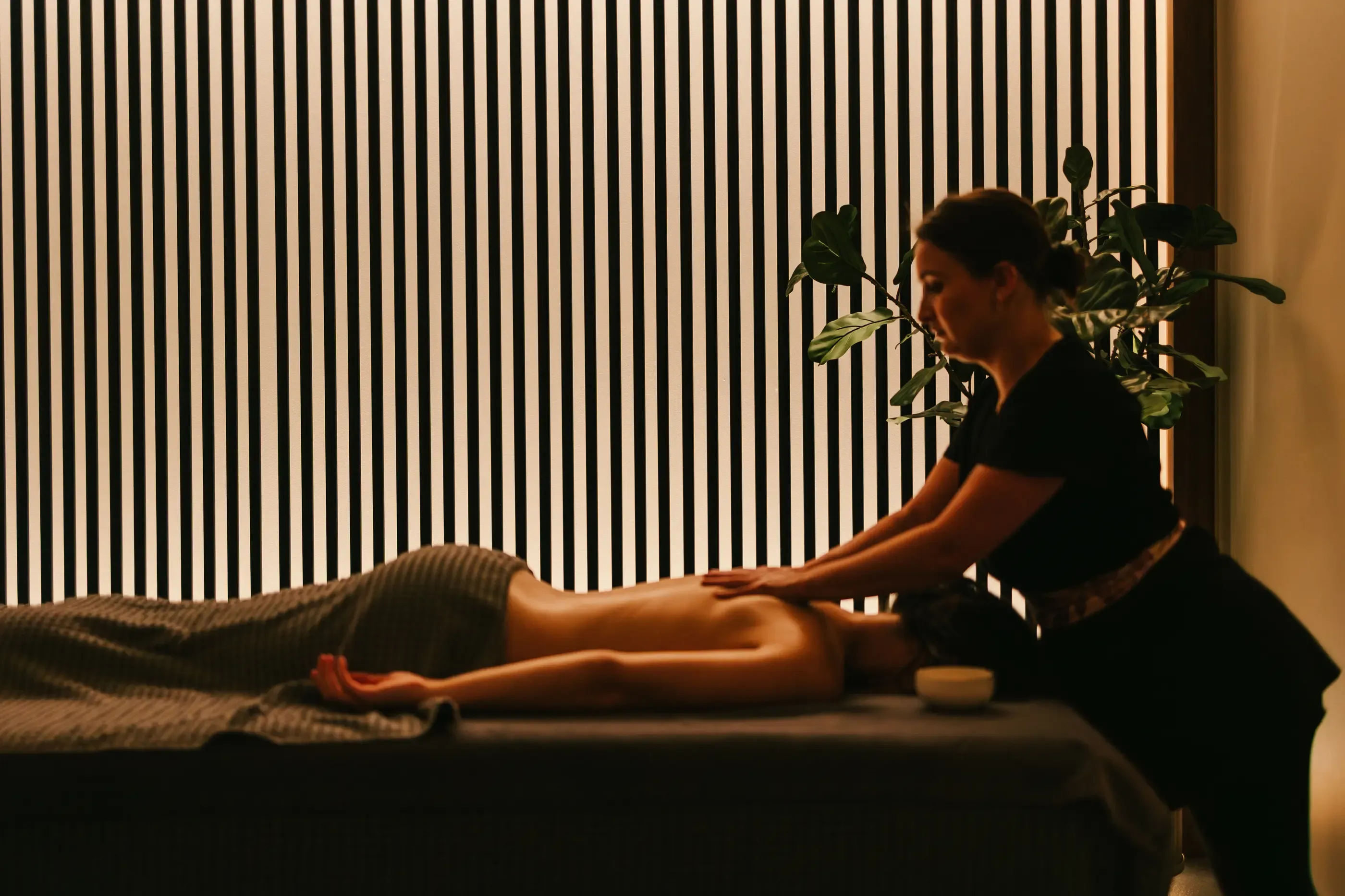 Une personne reçoit un massage sur une table dans une pièce avec une plante et des stores verticaux